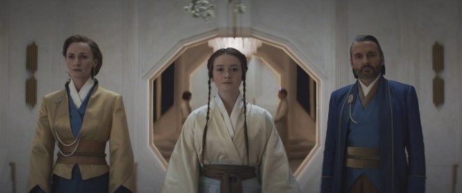 Three characters standing in a minimalist, elegant interior space with a geometric archway in the background. The central character, dressed in a light robe, is flanked by two figures in formal attire, each wearing different colored outfits.