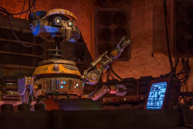 A robotic character with a round head and striped cap is seated at a control panel, interacting with a computer screen and surrounded by electronic equipment.